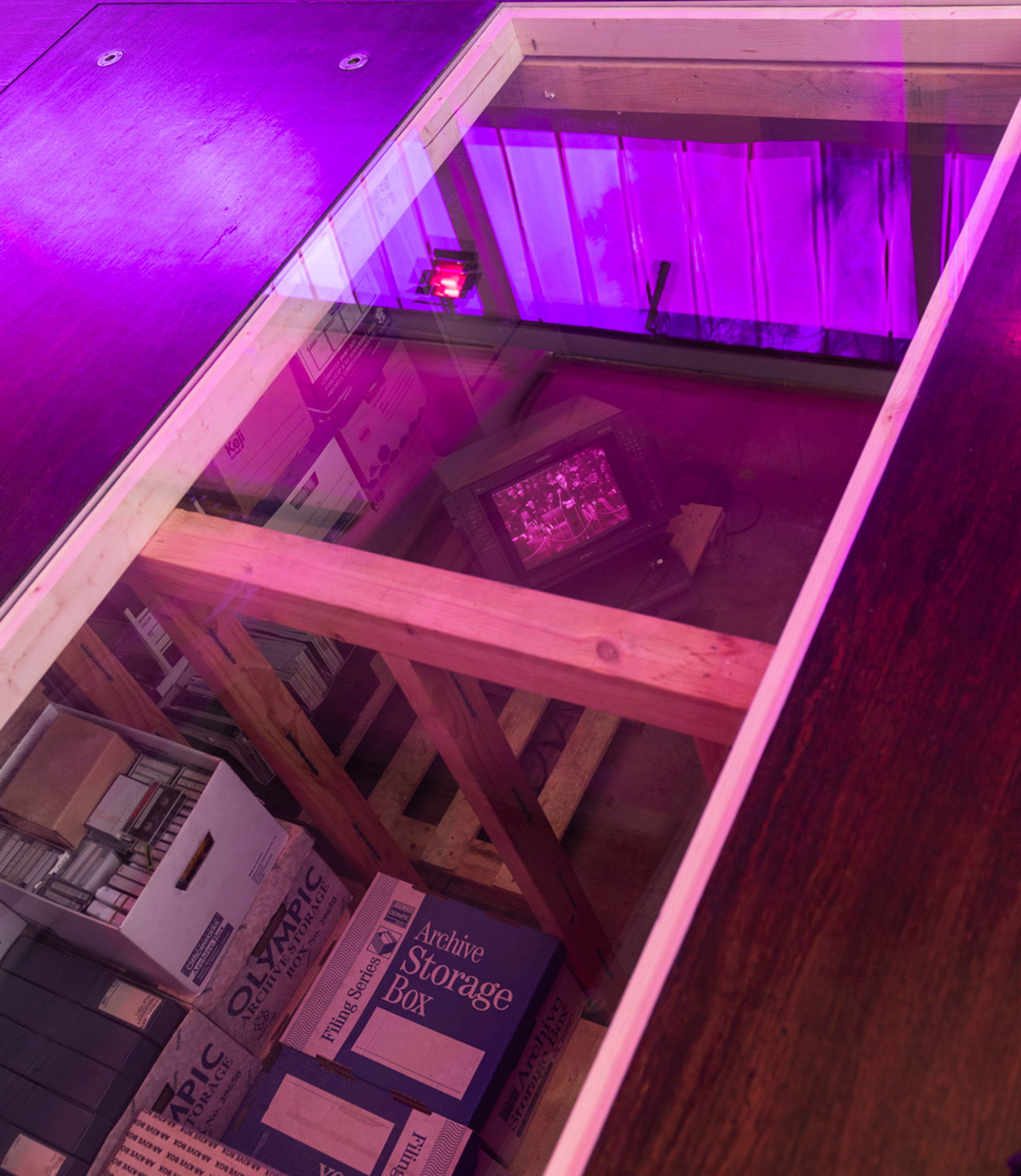 View through a glass-covered opening in the gallery floor into an archive storage area containing timber supports, document boxes, shelves, and a small vintage Sony broadcast monitor playing black-and-white footage, overlaid with reflections of purple windows and lights.