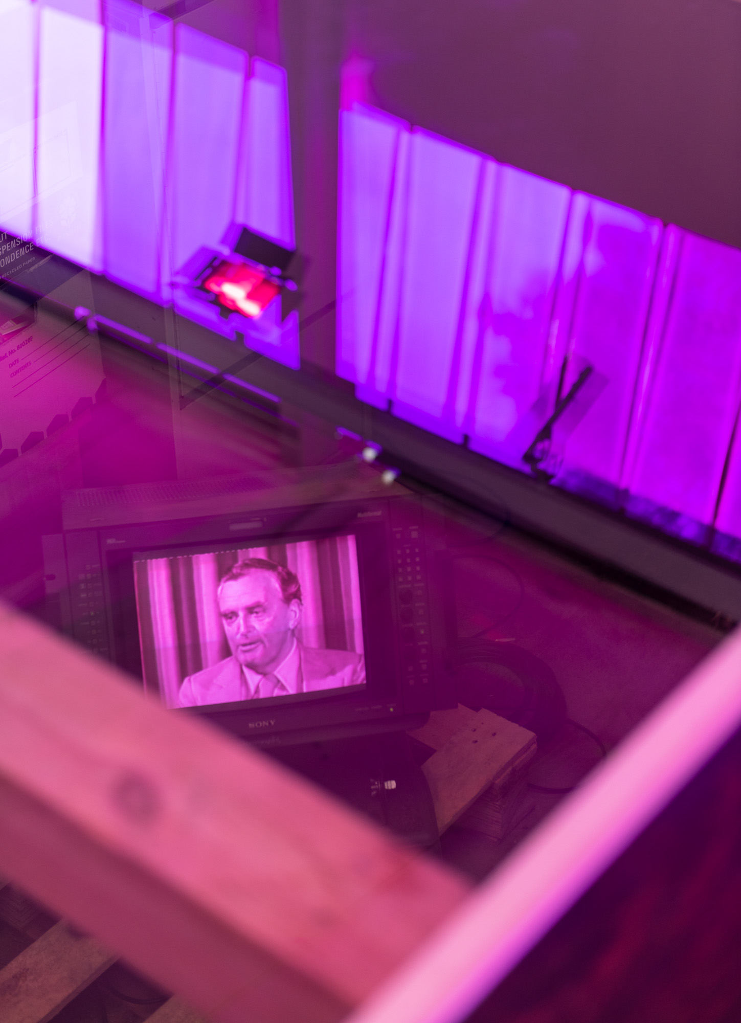 Close view through the glass floor into the archive below, where a vintage Sony broadcast monitor displays black-and-white footage of a man-Johannes Bjelke-Petersen- speaking, surrounded by papers, storage boxes, and vivid purple reflections from long purple windows.