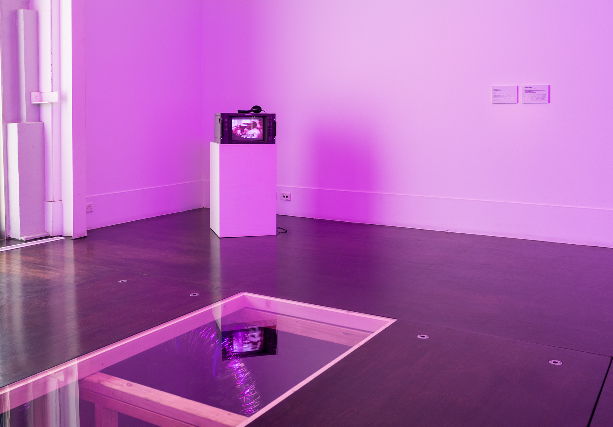 Installation view of a vintage Sony broadcast monitor with headphones on a white plinth, positioned beyond a glass-covered opening in the floor. The monitor and purple gallery lighting are reflected across the glass, with purple light filling the gallery.