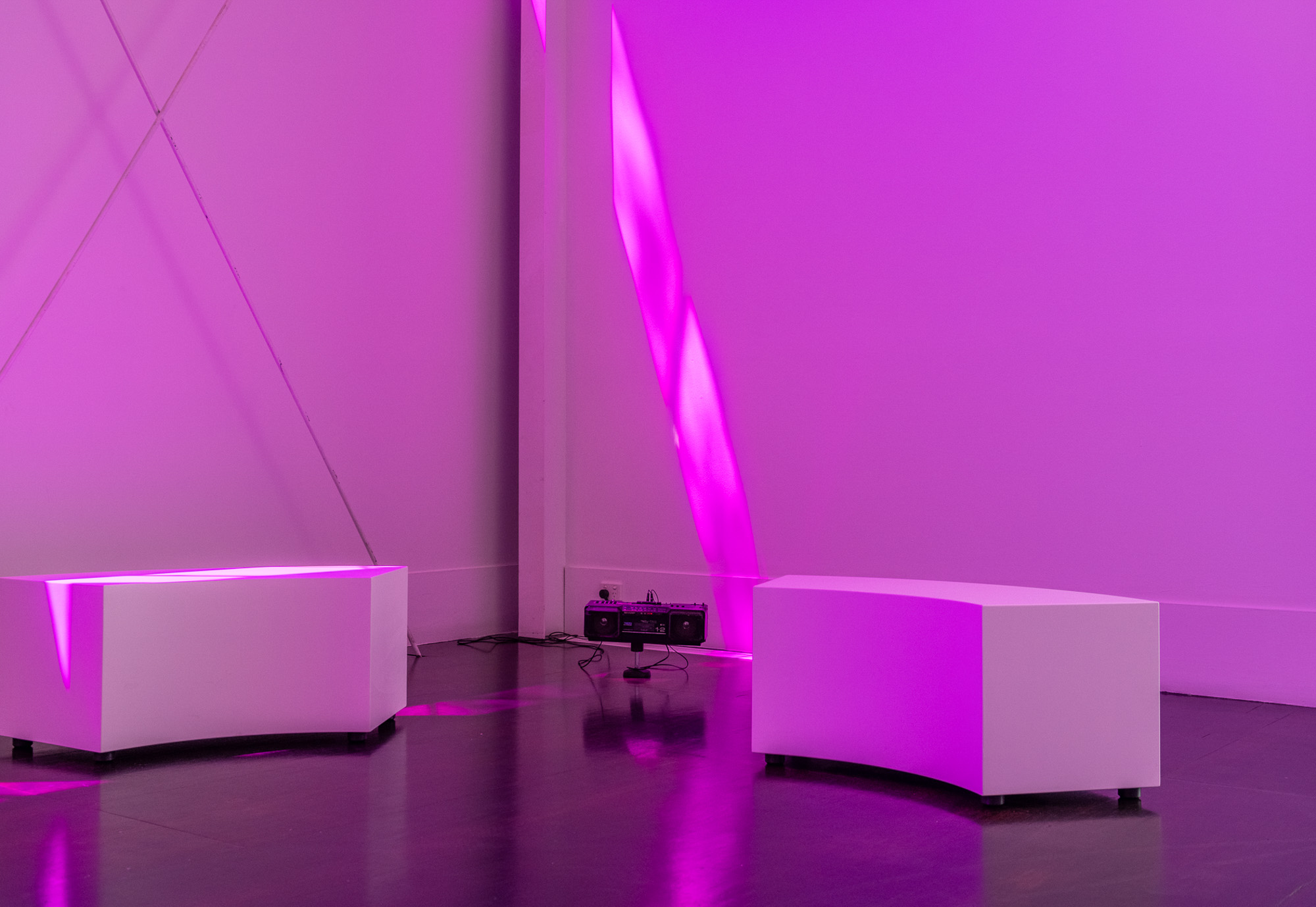 Installation detail of two curved white listening benches facing a black vintage portable radio and cassette player on the gallery floor, while vivid magenta light forms diagonal bands across the wall and reflects on the polished floor.