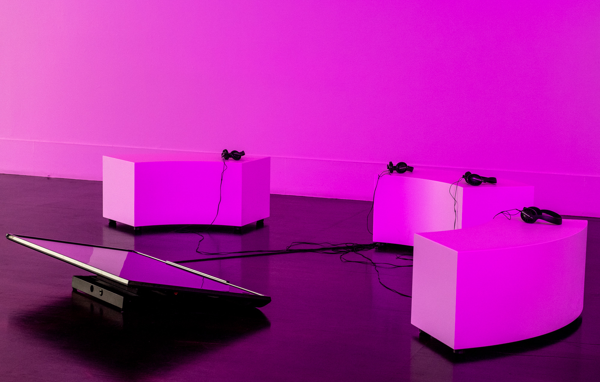 Three curved white listening benches arranged around a low, tilted video flatscreen television, with four black headphones, their cables resting across the seats and dark wood gallery floor. Intense magenta light fills the room and reflects upon the benches.