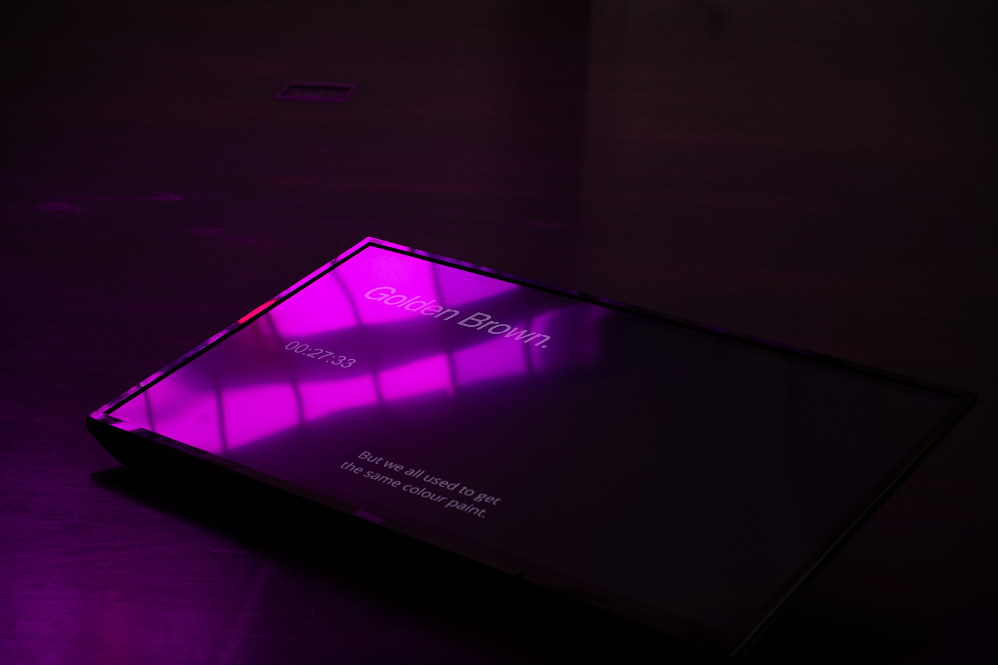 Low video screen displaying the name “Golden Brown,” a timestamp reading “00:27:33,” and the subtitle “But we all used to get the same colour paint,” beneath reflections of purple window light starkly contrast against the dark wood floor.