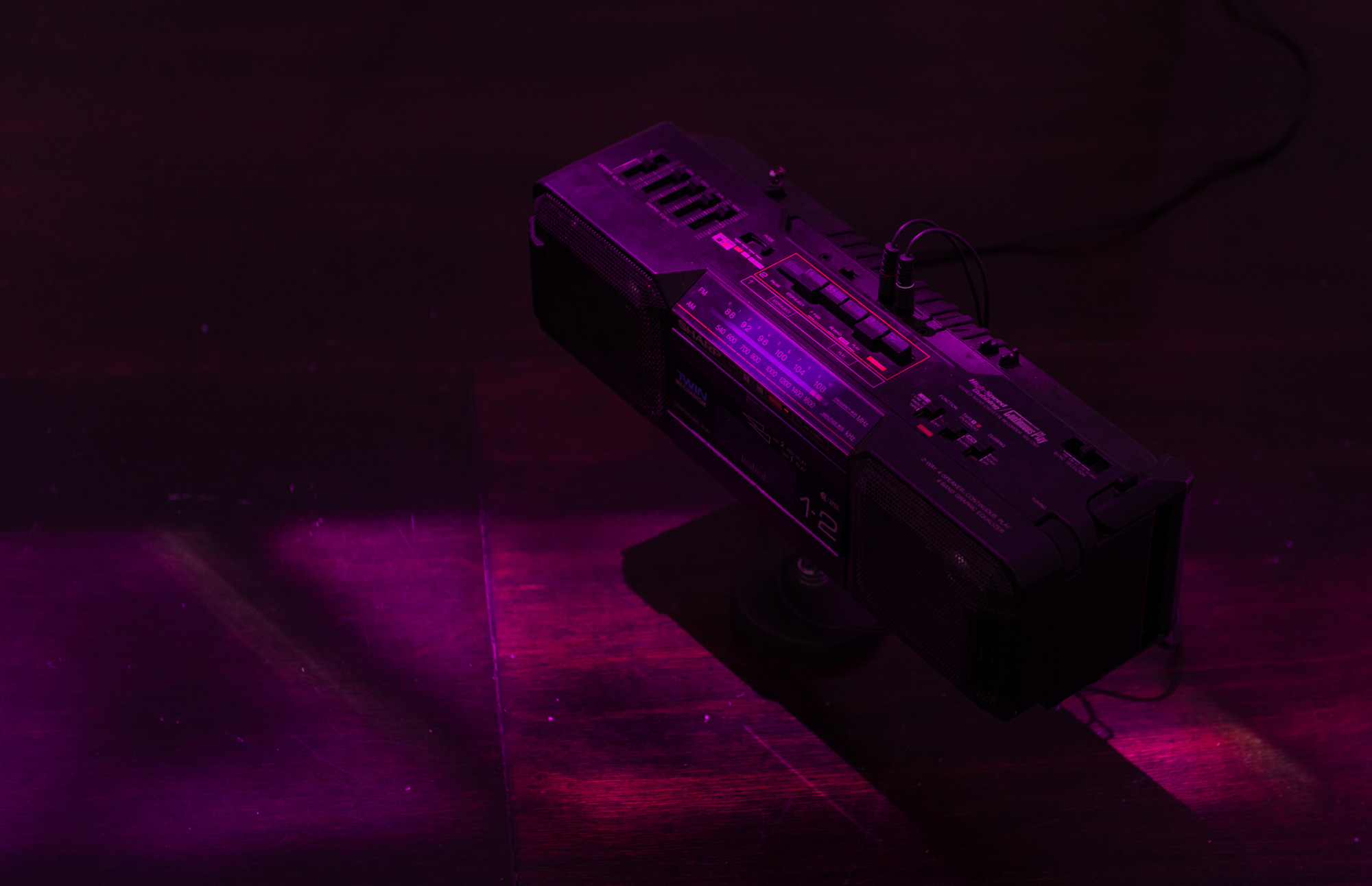 A vintage black portable radio and dual-cassette player positioned directly on the dark gallery floor, connected by audio cables and illuminated by concentrated magenta light.
