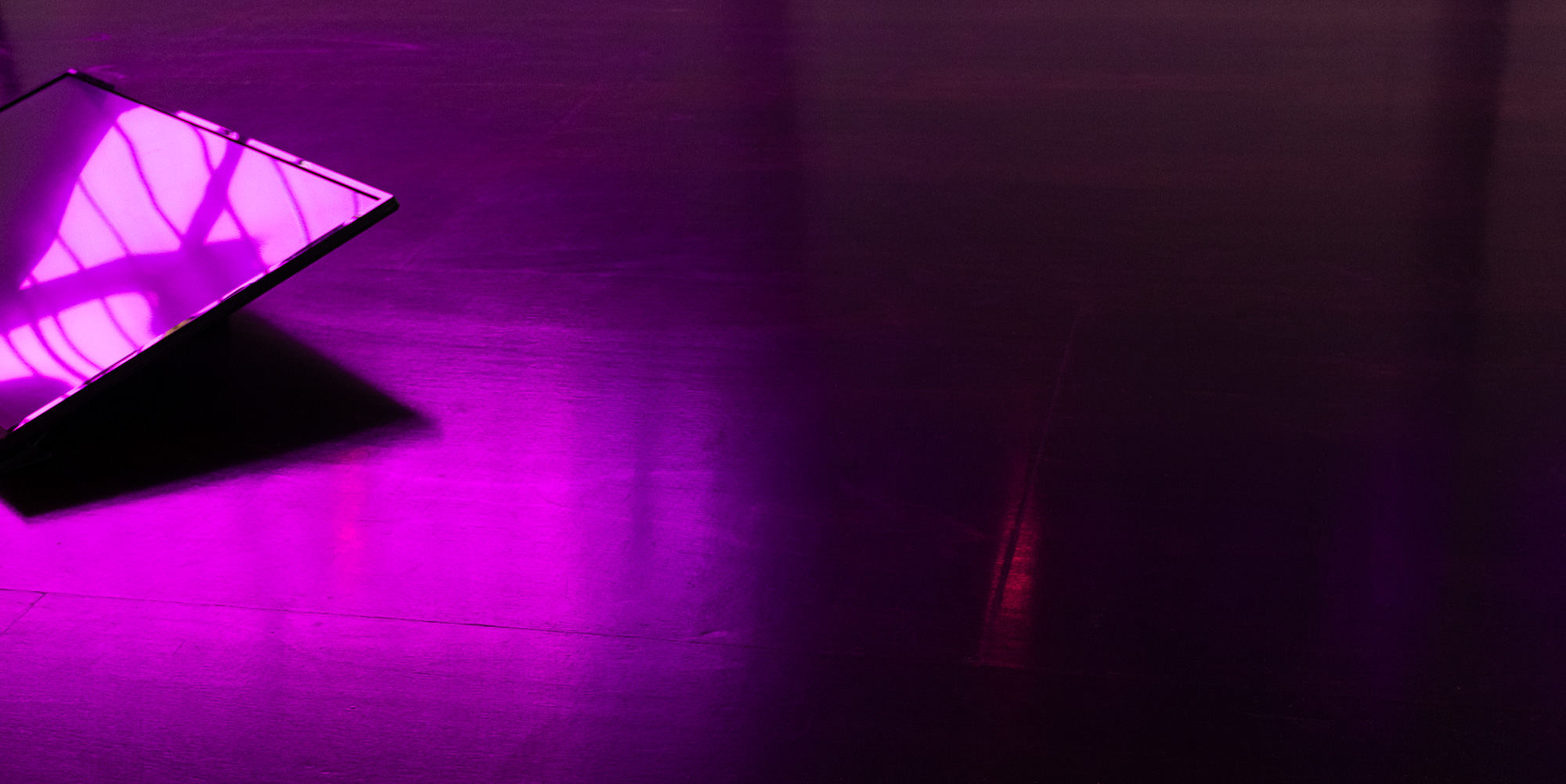 Detail of a low, tilted screen at the edge of the frame, its glossy surface reflecting the purple gallery windows and casting a deep angular shadow across the dark wooden floor.