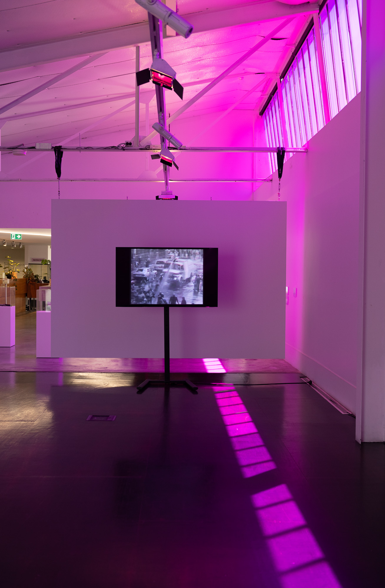 Flatscreen monitor on a tall stand shows black-and-white archival footage of a water cannon spraying into a crowded street, positioned in front of a suspended white wall, as purple-filtered sunlight fills the gallery space and forms a long grid across the gallery floor.