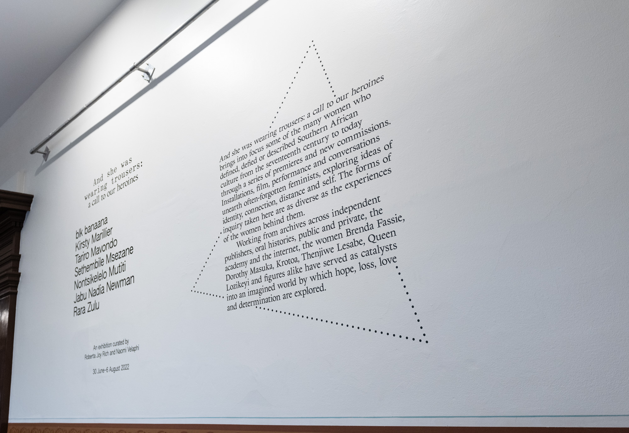 Exhibition wall text for "And she was wearing trousers: a call to our heroines", listing artists blk banaana, Kirsty Marillier, Tariro Mavondo, Sethembile Msezane, Nontsikelelo Mutiti, Jabu Nadia Newman and Rara Zulu, curated by Roberta Joy Rich and Naomi Velaphi, 30 June–6 August 2022. The introductory text is set on a slant inside a dotted triangle.