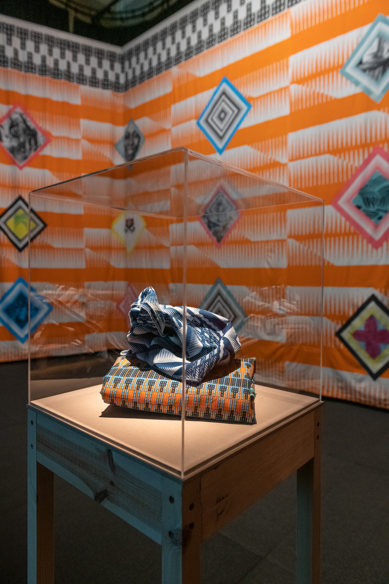 A bunched indigo-and-white cloth resting on a folded orange geometric print inside a clear acrylic case, with the orange patterned fabric walls blurred behind.