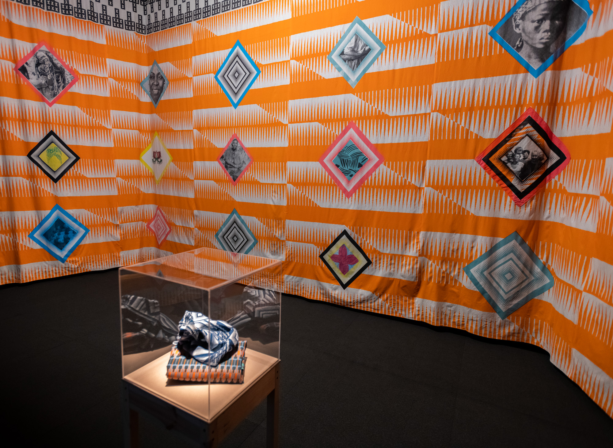 Corner view of the orange-and-white textile installation walls, decorated with photo portraits of African women and geometric motifs in colourful diamond frames. A clear case holding folded printed fabrics sits on a stand in front.