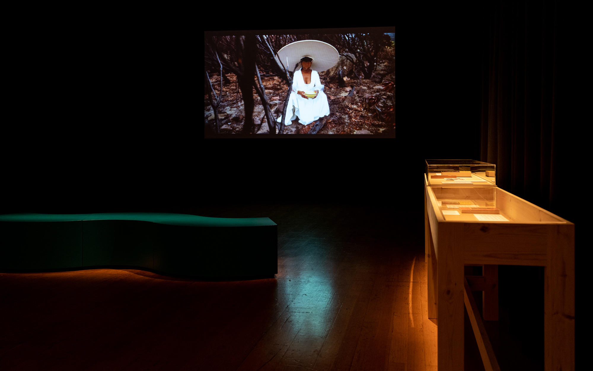 Video projection of an African woman in a flowing white dress and wide circular hat, seated among burnt shrubs holding a yellow bowl. A teal S-shaped bench and a timber vitrine face the screen in the dark room.