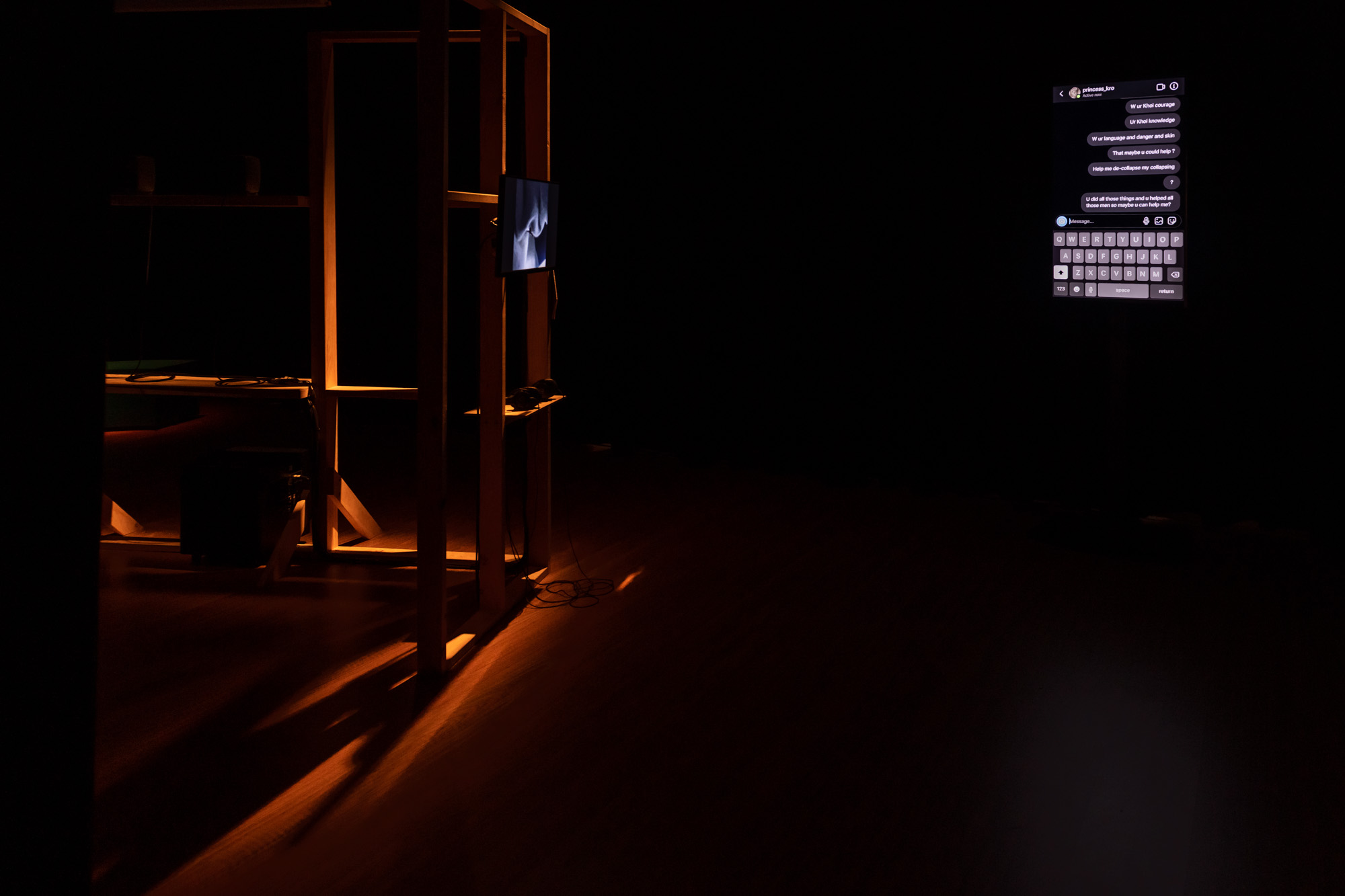 A dark gallery with a vertical screen displaying an Instagram chat with "princess_kro", with messages including "W ur Khoi courage" and "U did all those things and u helped all those men so maybe u can help me?". At left, a timber frame structure holds a small glowing screen.