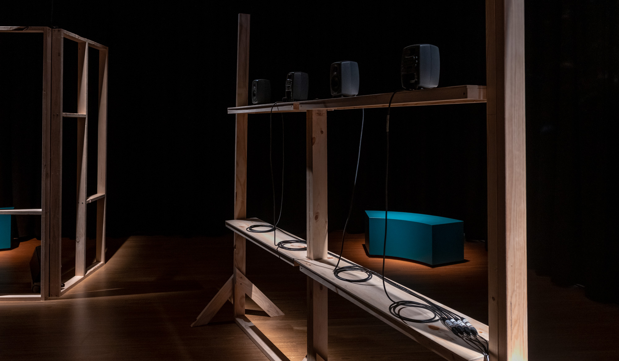 Detail of the timber frame shelving holding four small grey speakers with neatly coiled cables, spot lit along the wood frame. A teal curved bench sits in the darkness beyond.