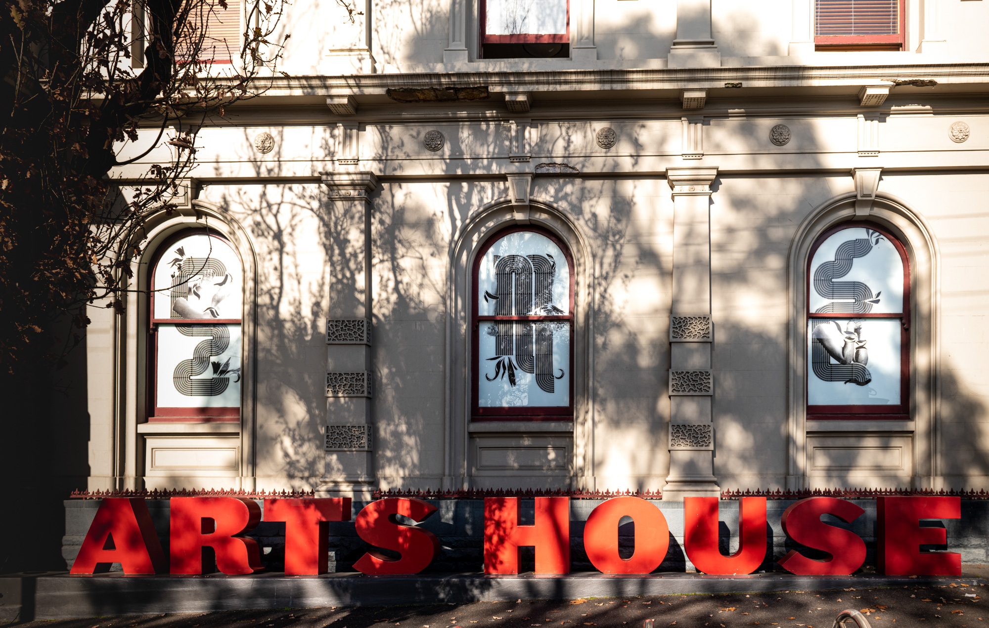 The heritage facade of Arts House, North Melbourne, with giant red three-dimensional letters spelling "ARTS HOUSE" along the base. Three arched windows hold black-and-white vinyl artworks of woven African braid patterns and photographic body fragments.