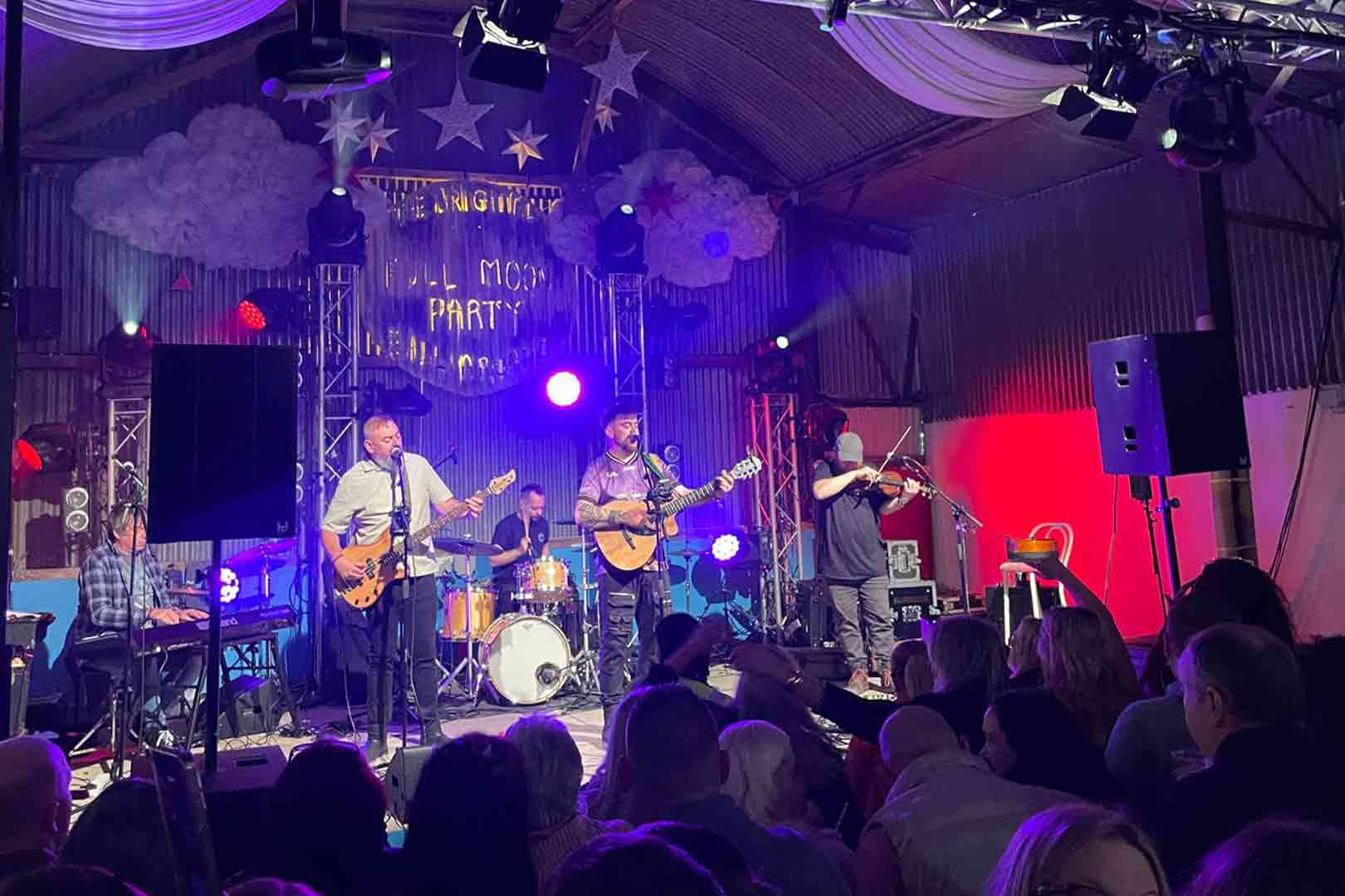 Band performing on stage with guitar, violin, keyboard, and drums during a lively indoor concert with an engaged audience.