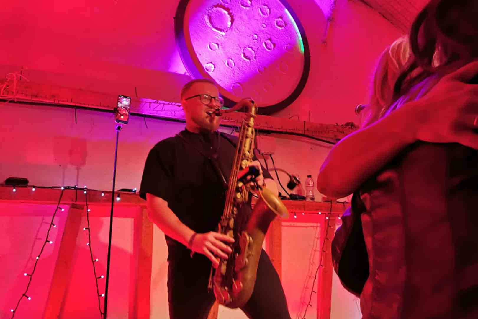 Musician playing saxophone under purple and red lighting with a large circular wall decoration behind.