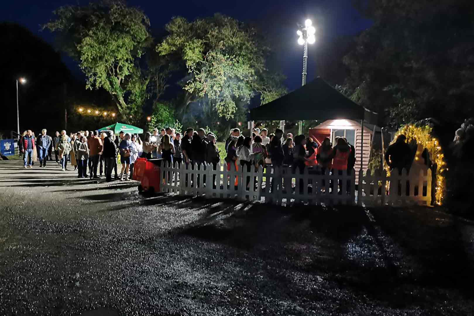A crowd of people gathered at a nighttime outdoor event near a lit tent and a white picket fence, with trees and string lights in the background.
