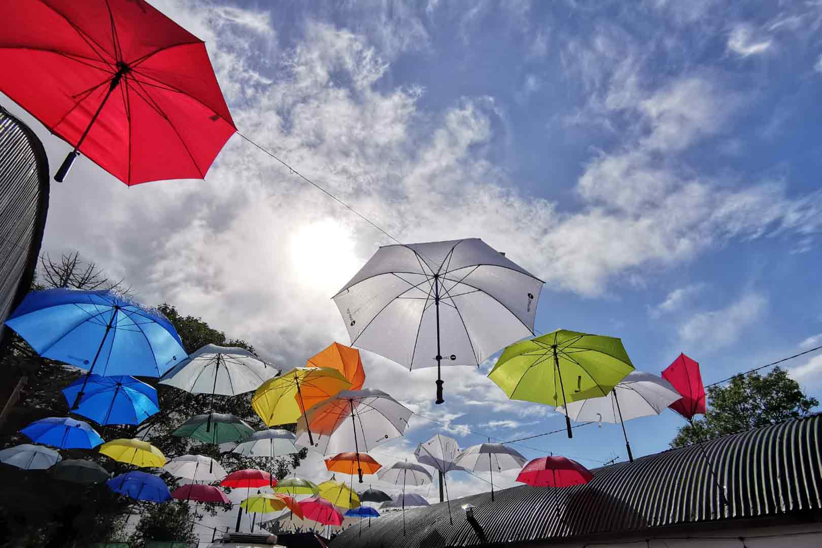 Colorful umbrellas suspended on wires against a partly cloudy sky with the sun shining.