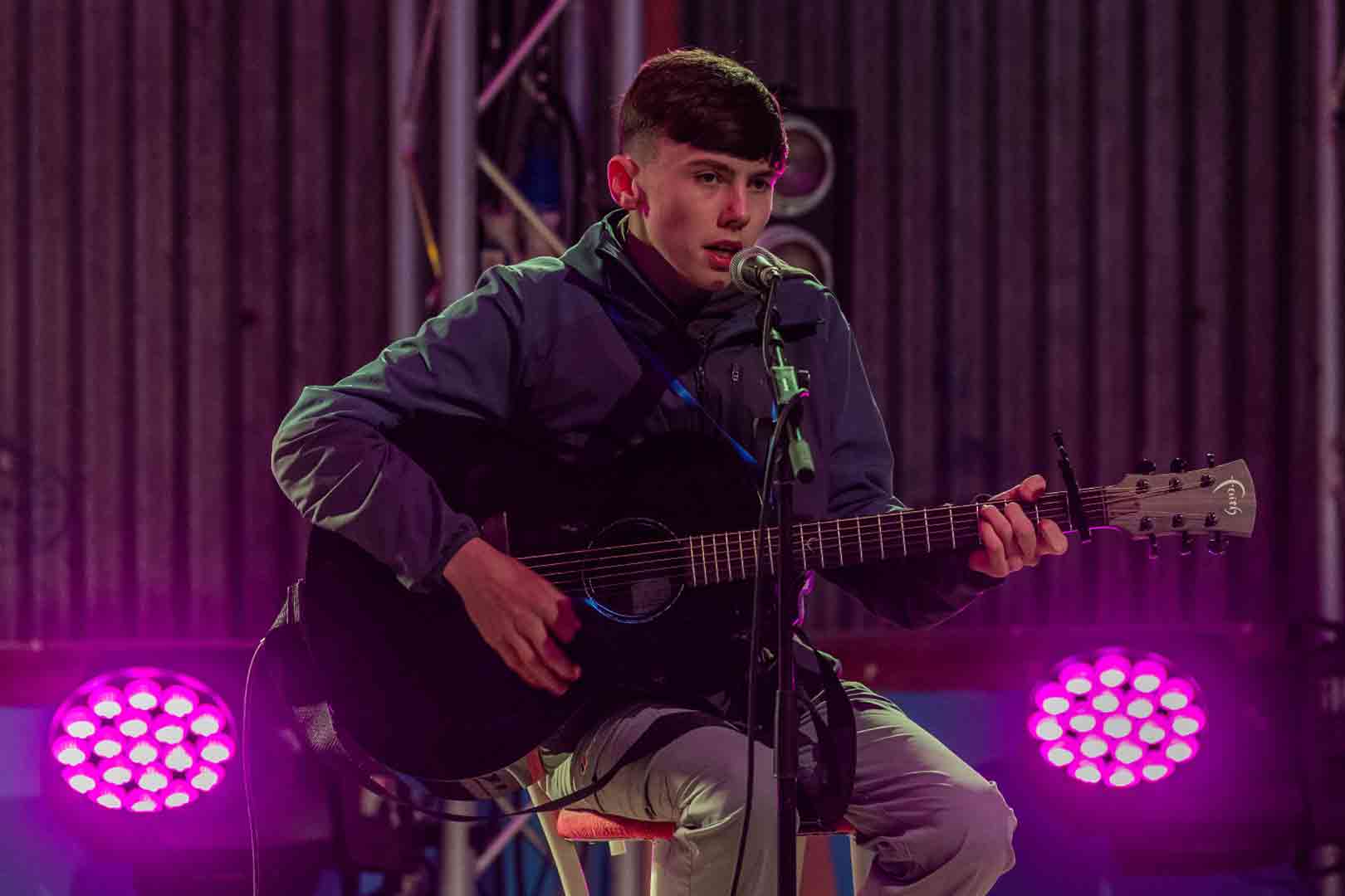 Young man playing an acoustic guitar and singing into a microphone under purple stage lights.