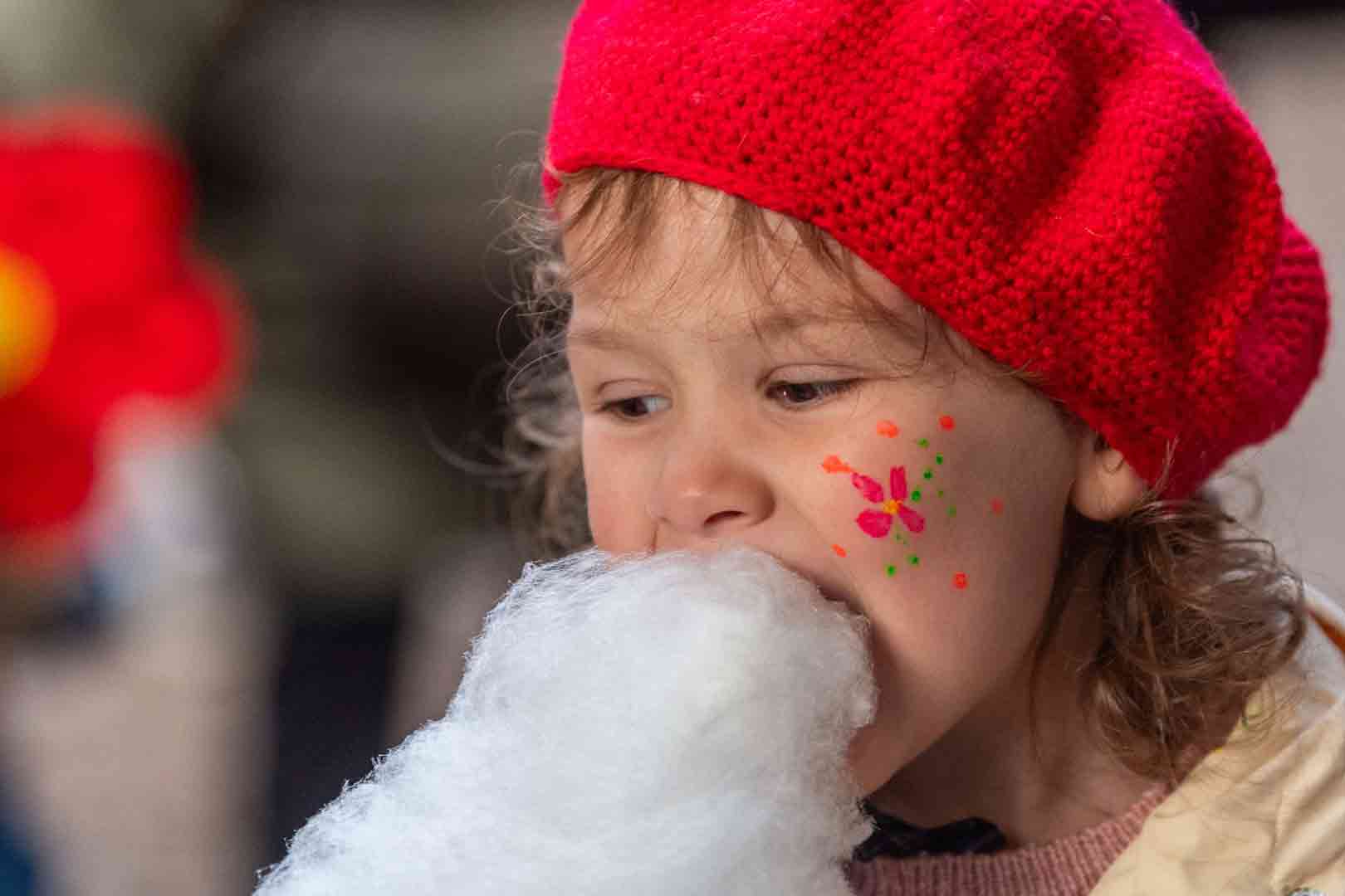 Child wearing a red knit hat eating white cotton candy with face paint of a colorful flower on the cheek.