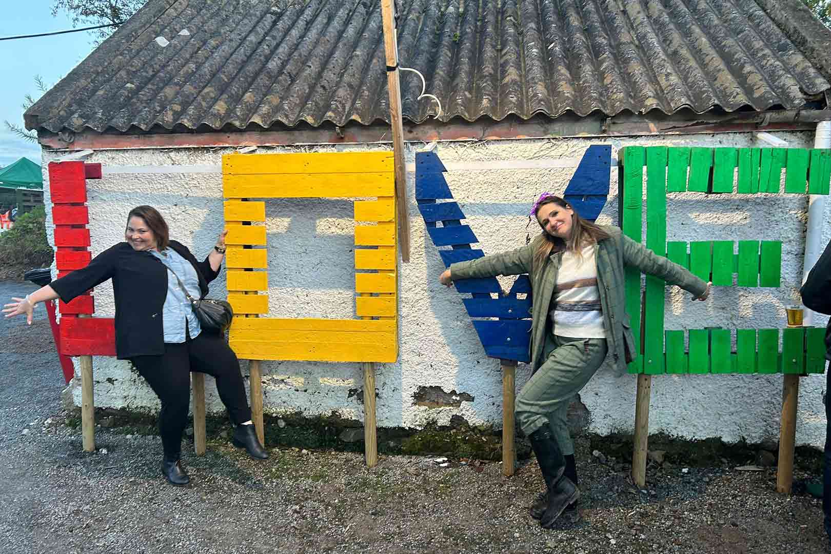 Two women posing joyfully with large colorful wooden letters spelling LOVE against a white textured wall.