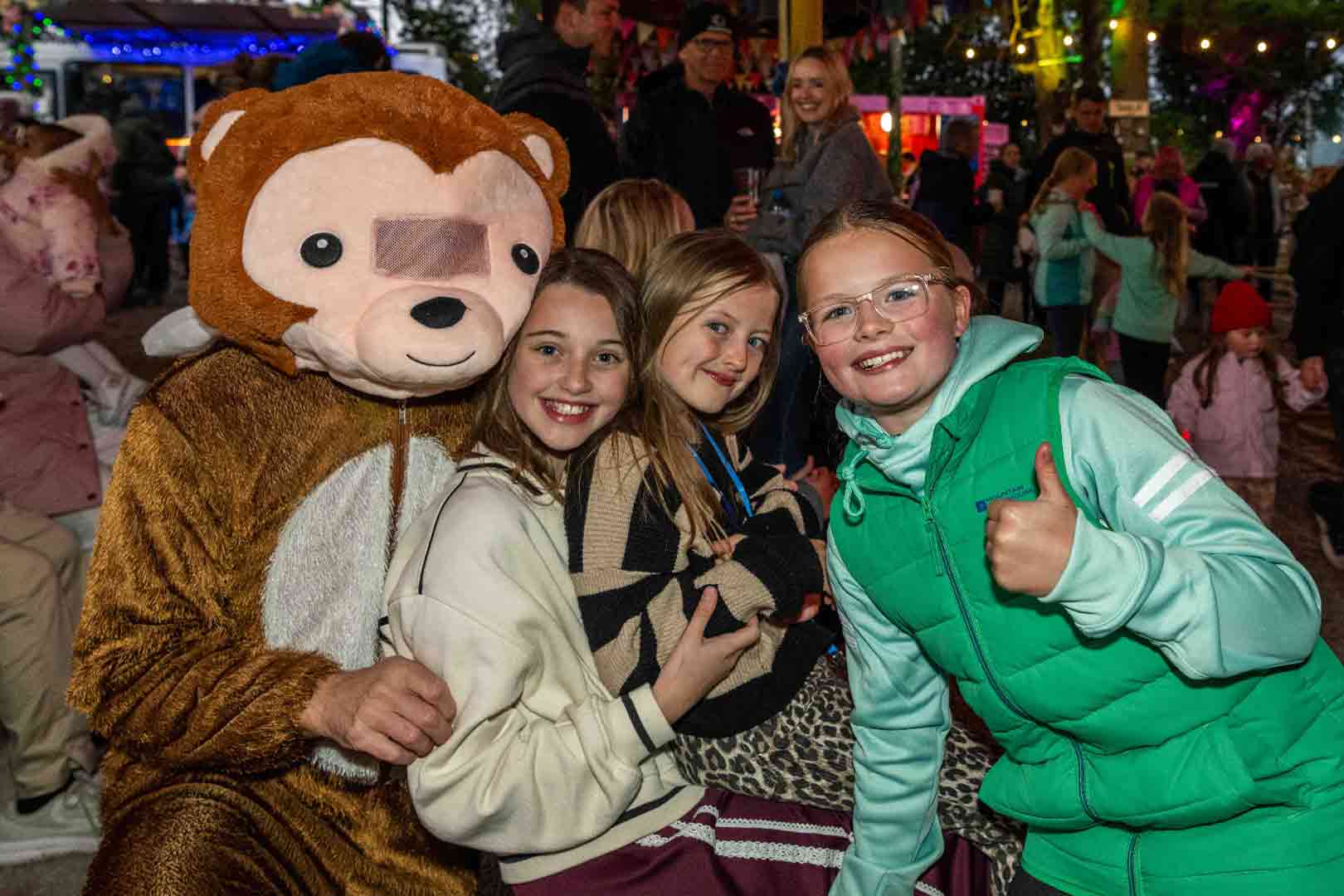 Three smiling children posing with a person dressed in a brown monkey costume at an outdoor event, with festive lights in the background.
