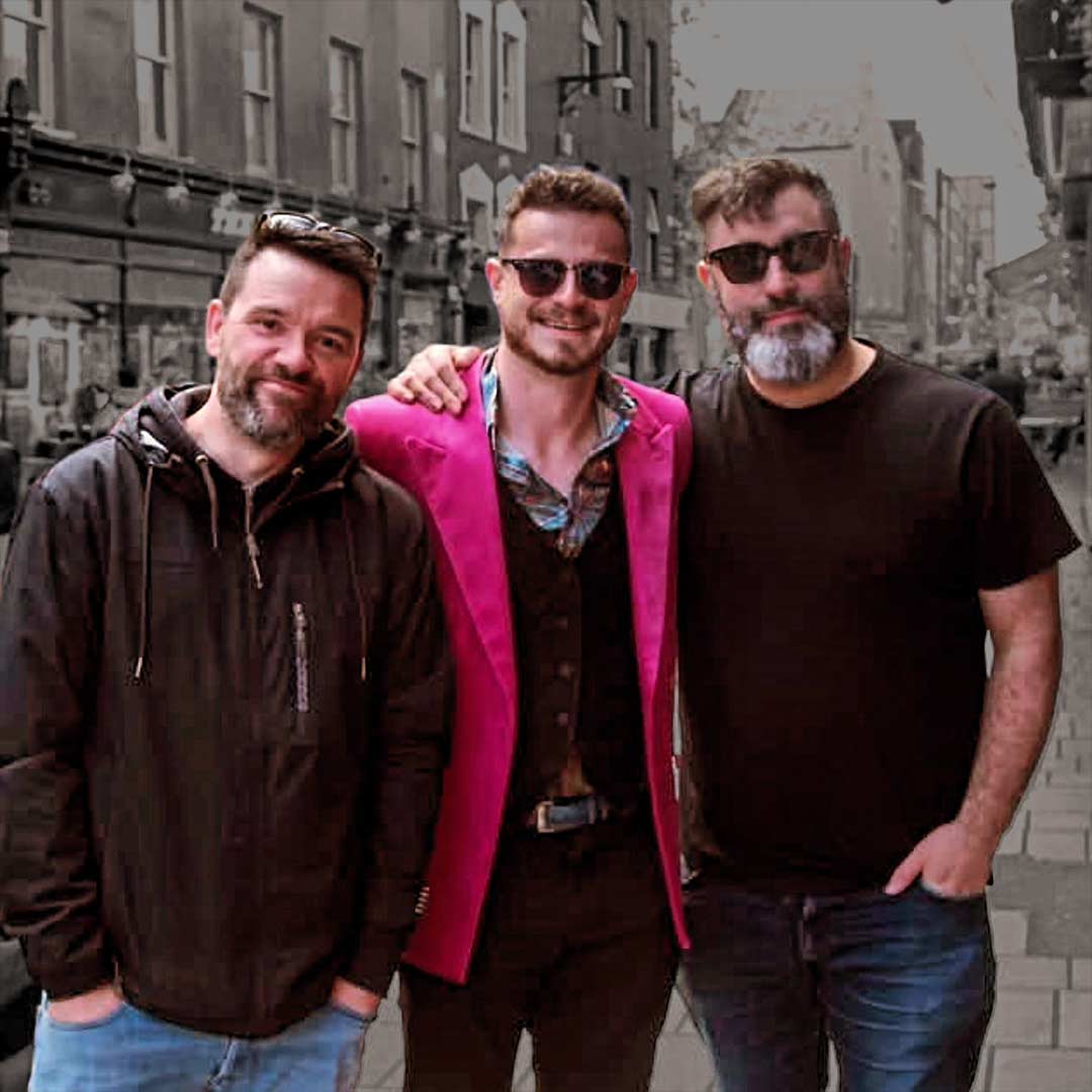 Three men standing side by side on a city street, two wearing sunglasses and casual clothes, the one in the middle wearing a bright pink blazer.
