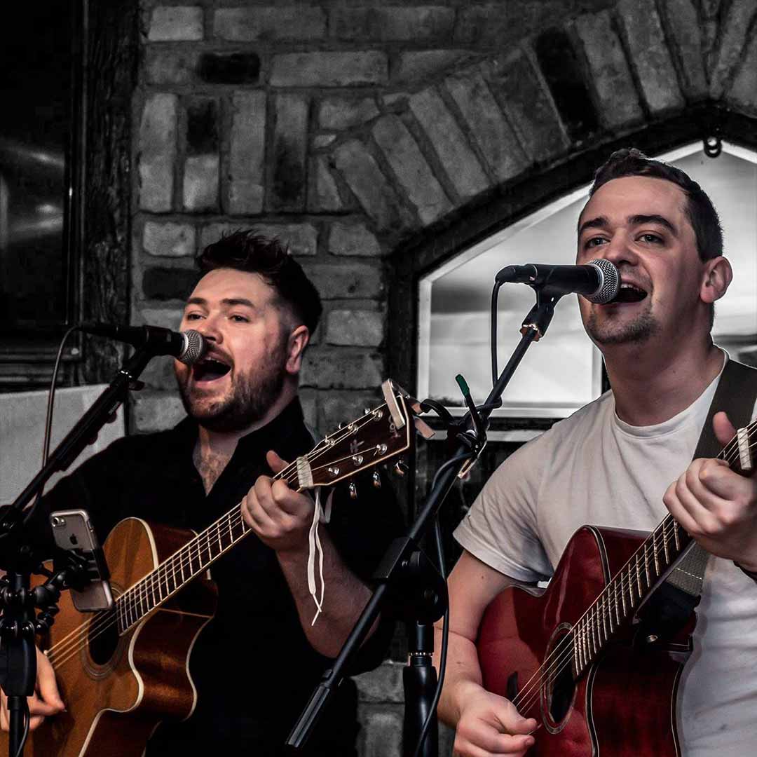 Two men singing into microphones and playing acoustic guitars in front of a stone wall.