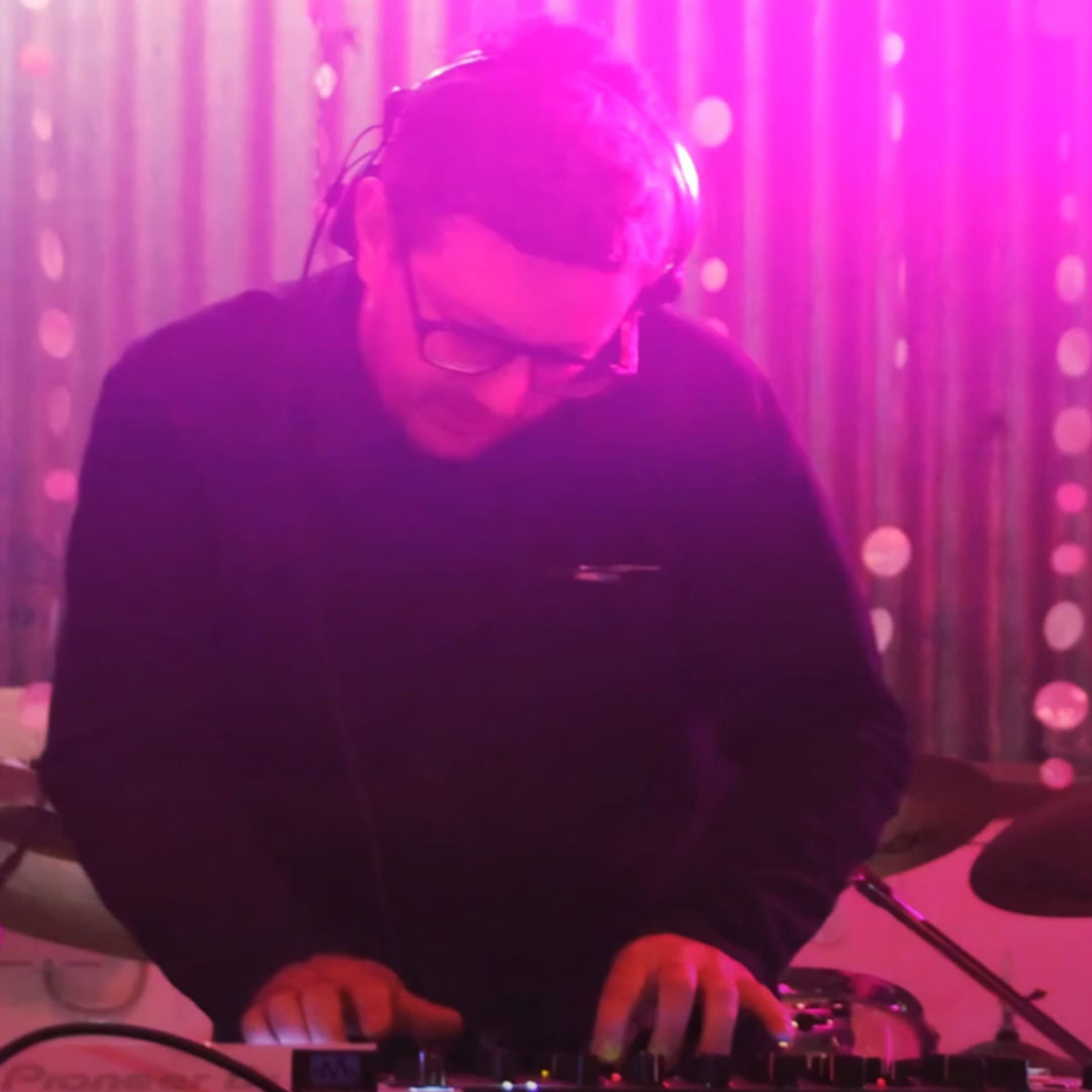 DJ with headphones and glasses mixing music on a console under pink stage lights.