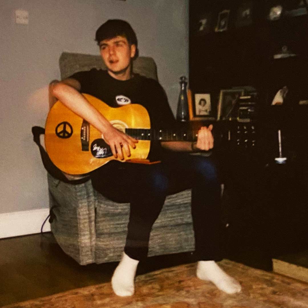 Young man sitting on a chair playing an acoustic guitar with peace symbol and stickers.