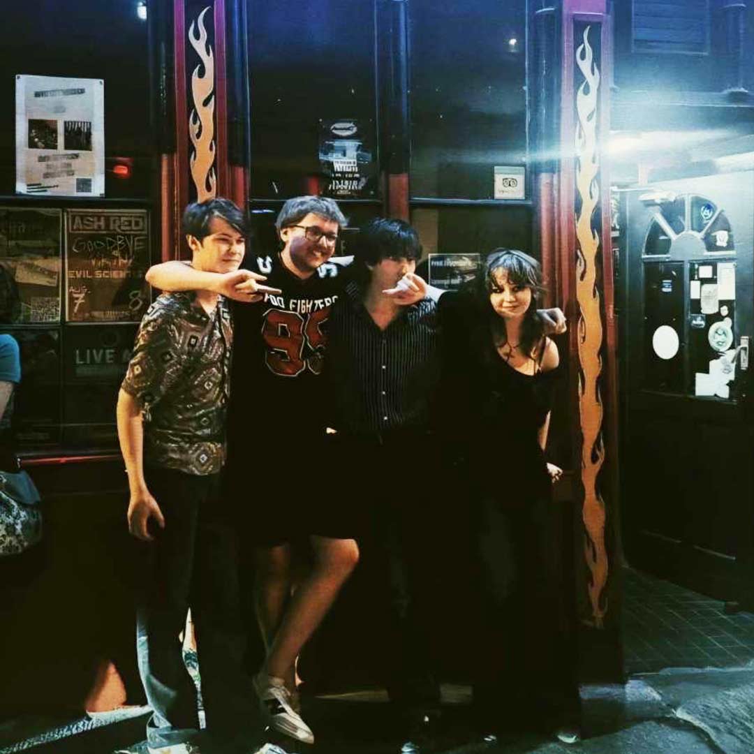 Four young people standing close together at night outside a venue with flame-decorated pillars.