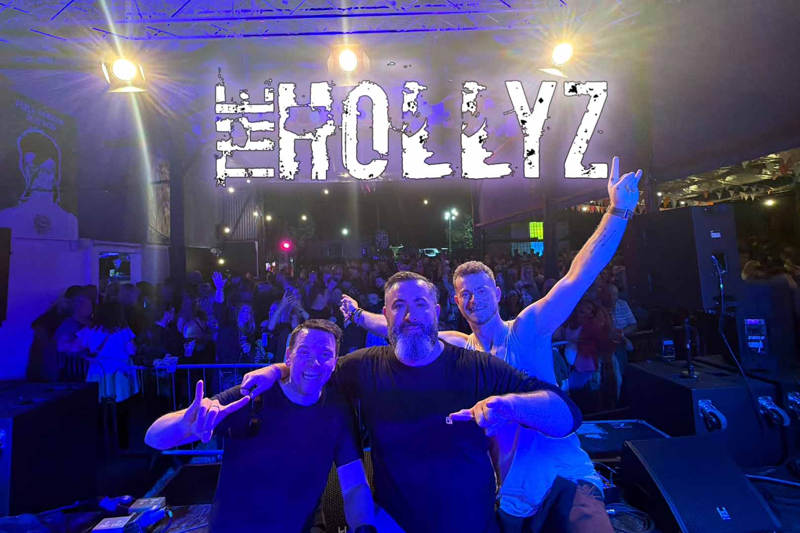 Three male performers on stage posing for a photo with their arms around each other and making rock hand gestures, with a crowd of people and stage lights behind them, and the text 'THE HOLLYZ' lit above them.