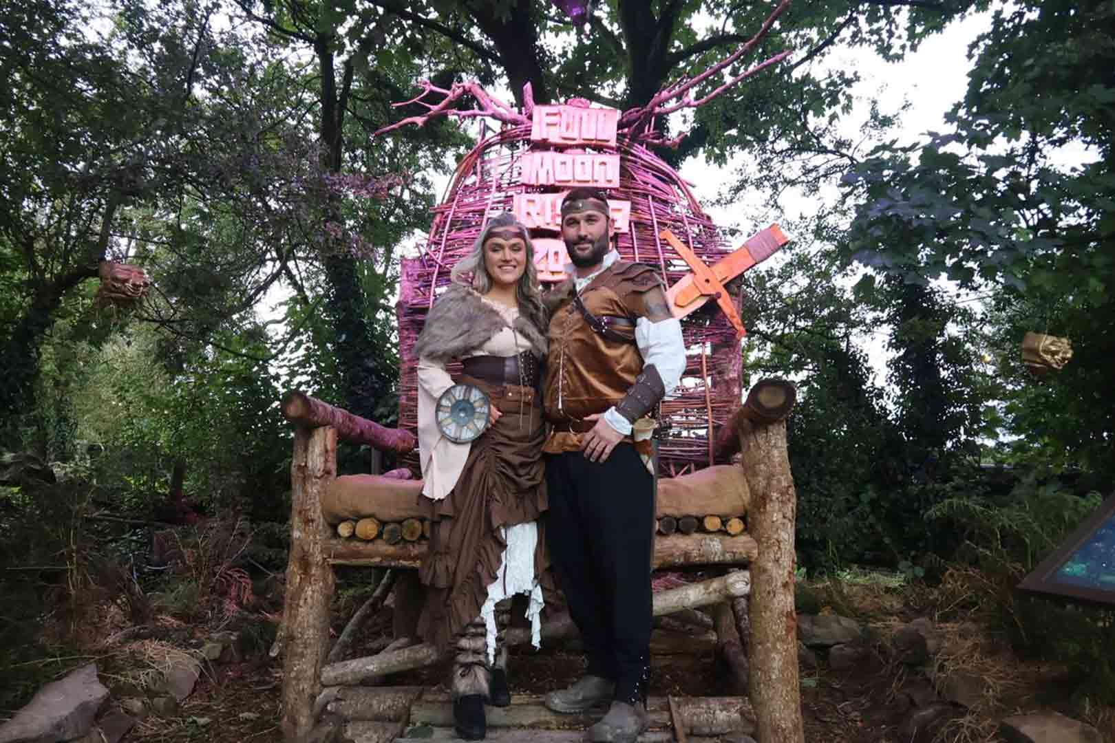 A man and woman dressed in medieval fantasy costumes standing on a rustic wooden bench outdoors with trees around and a purple woven structure with signs behind them.