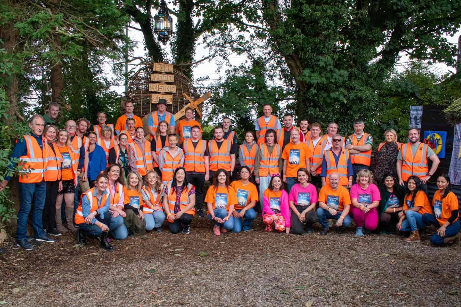 Large group of about 40 people posing outdoors among trees, most wearing orange safety vests and some wearing orange or pink t-shirts, with a rustic wooden sculpture and signs behind them.