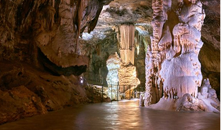 Illuminated cave interior featuring large stalactites and stalagmites with a pathway and railing.