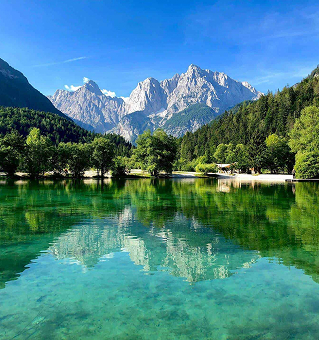 Clear lake with vibrant green reflection, surrounded by trees and mountains under a bright blue sky.
