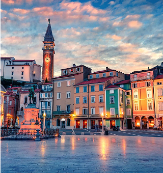 Evening view of a historic European town square with colorful buildings, a clock tower, and a statue, under a dramatic cloudy sky.