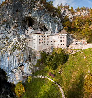 Medieval castle built into a rocky cliff surrounded by autumn foliage and a winding path below.