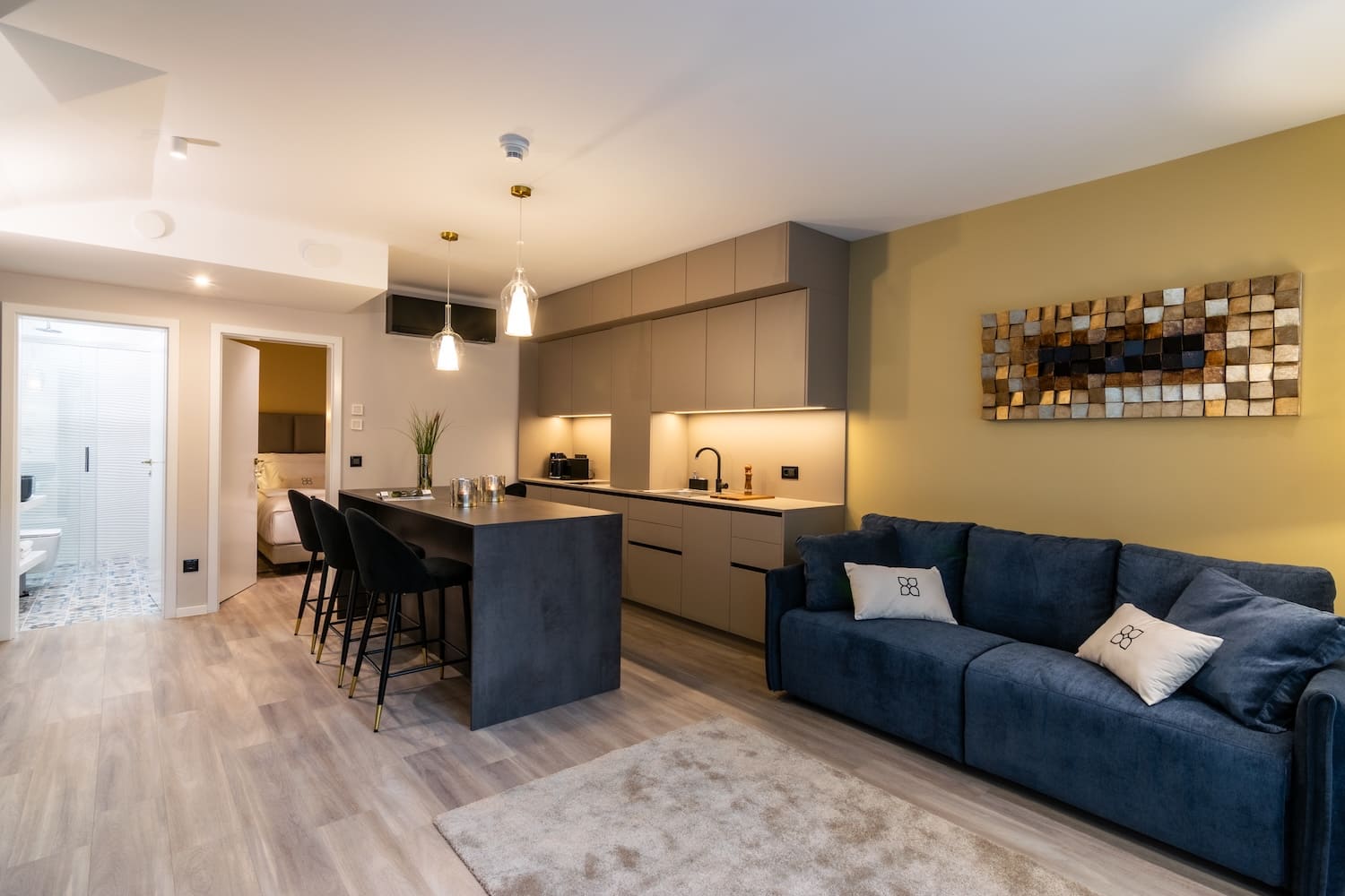 Modern living space with a blue sofa, beige kitchen cabinetry, island with black bar stools, and a doorway leading to a bedroom and bathroom.