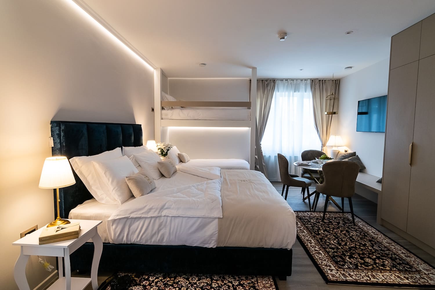 Modern hotel room with a large bed, bunk bed, small dining table with chairs, and a wall-mounted TV.
