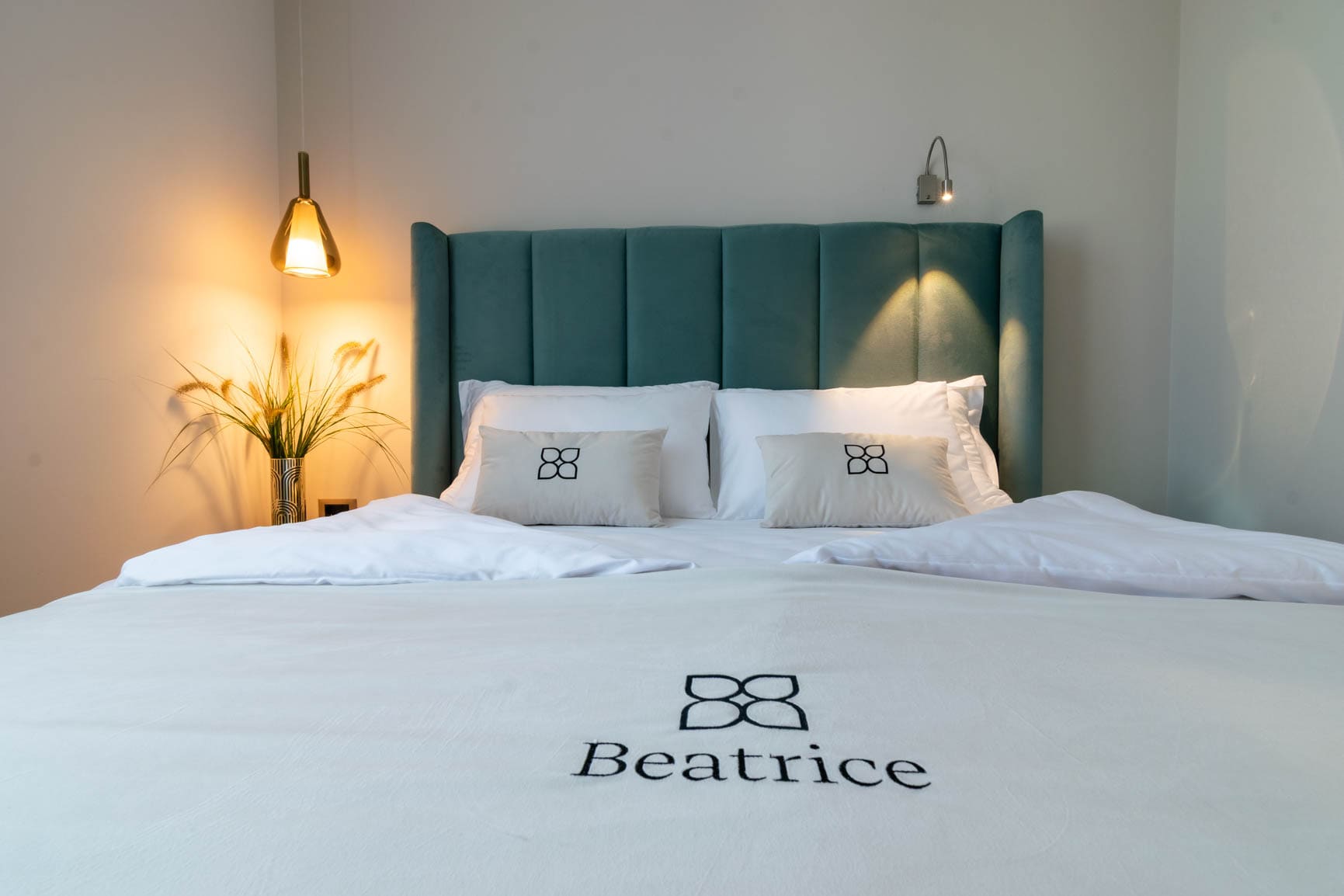 Modern bed with teal upholstered headboard, white bedding embroidered with 'Beatrice,' lit by two wall lamps and a vase with wheat stalks.