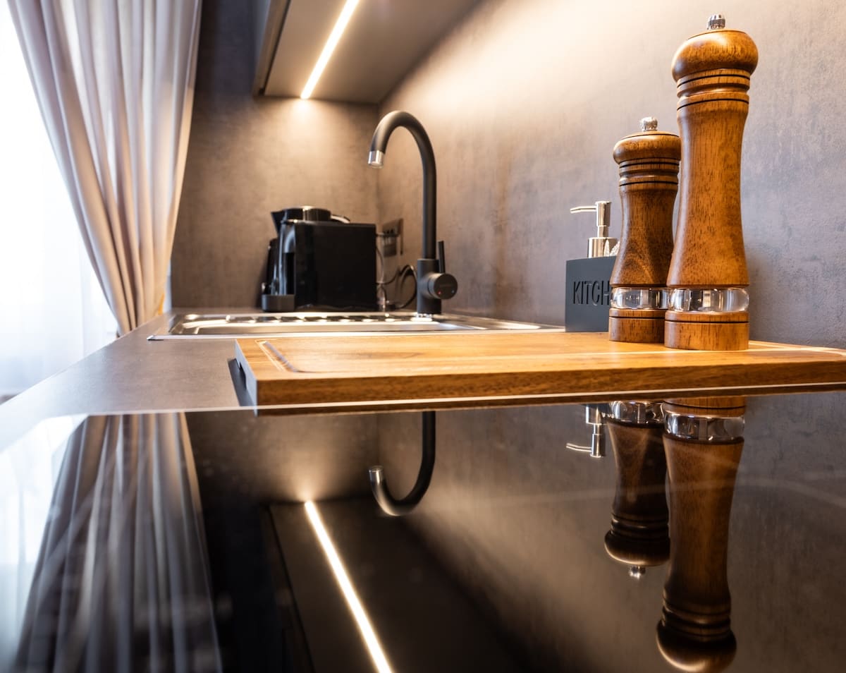 Modern kitchen countertop with wooden cutting board, salt and pepper grinders, black faucet, coffee machine, and under-cabinet lighting.