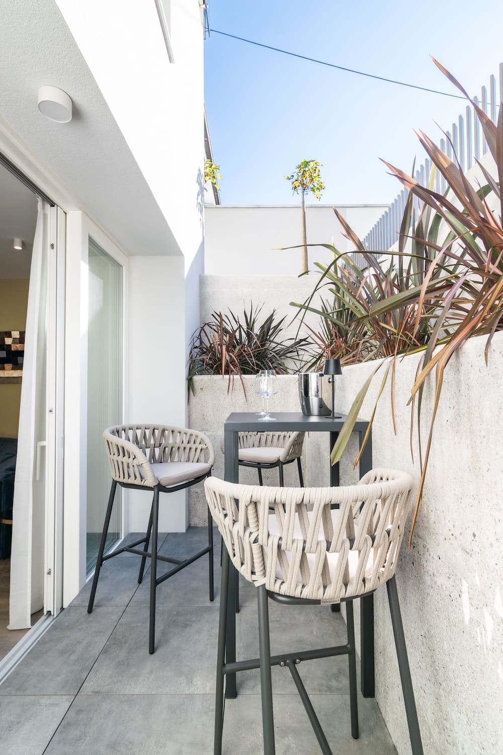 Small outdoor patio with woven beige chairs and a black table with a wine glass and ice bucket, next to a white building and plants.