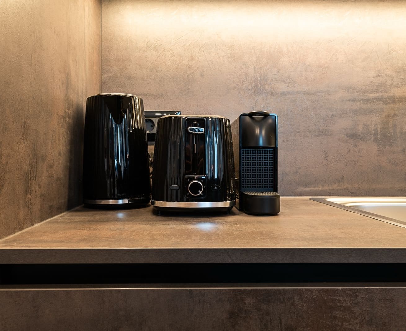 Black electric kettle, black toaster, and black capsule coffee machine on a brown kitchen countertop.
