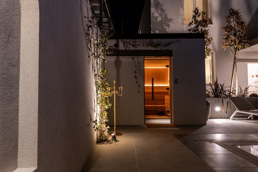 Evening view of a modern outdoor patio with a lit sauna room, potted plants, and a poolside lounge chair.