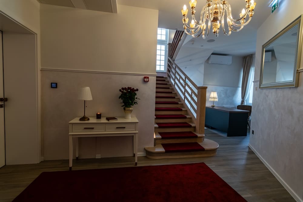Indoor space with stairs covered by a red carpet runner, a white console table with a lamp, candle, and flower vase, a red rug, chandelier, and a room with desk and lamp in the background.