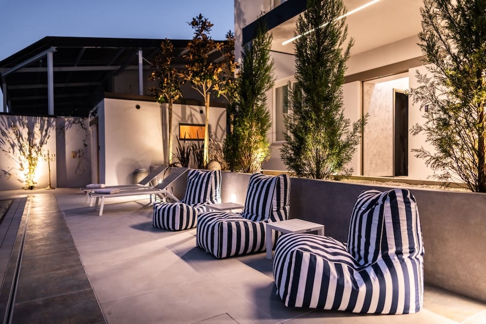 Modern outdoor patio at dusk with striped bean bag chairs, white side tables, and lounge chairs beside a building with illuminated trees.