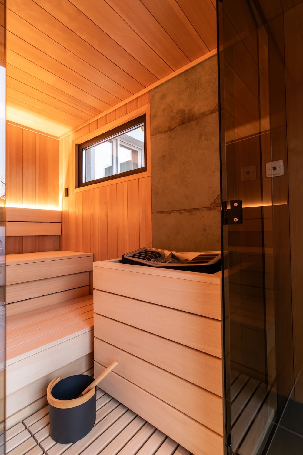 Modern wooden sauna interior with bench seating, a heater with stones, a small window, and a black bucket with a wooden handle on the wooden floor.