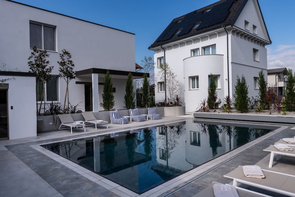 Modern white house with solar panels and a rectangular swimming pool surrounded by lounge chairs and greenery.