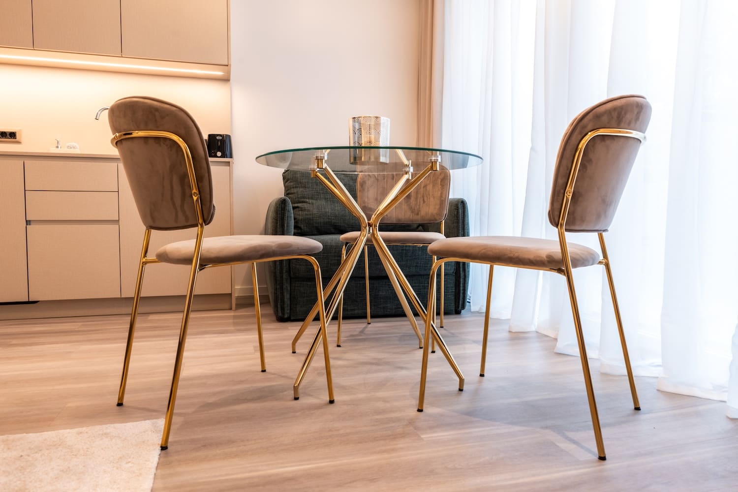 Modern dining area with a round glass table with gold legs, two beige velvet chairs with gold frames, and a dark upholstered armchair in the background.