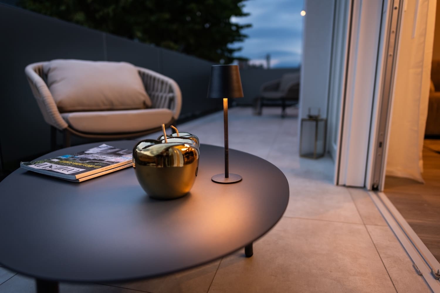 Outdoor patio table with a small modern lamp, gold decorative apples, and a magazine, with a cushioned chair and sliding glass door in the background during evening.