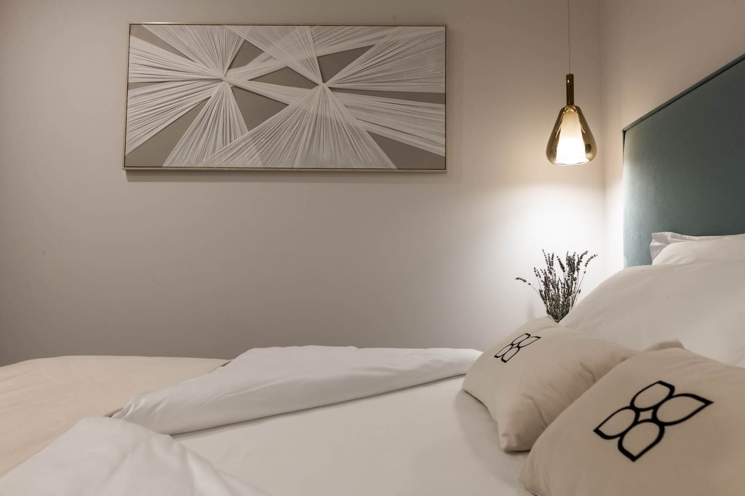 Cozy bedroom corner with a white bed, decorative pillows, a hanging lamp, and abstract wall art.